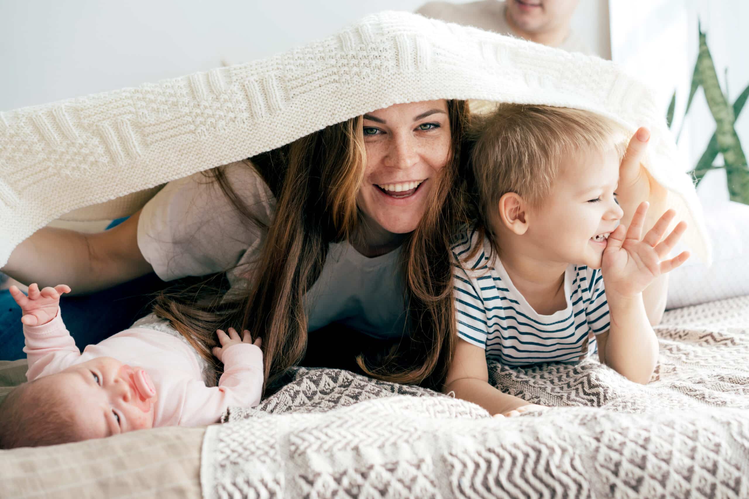 Happy mommy plays with the children on the bed and covers the children and herself with a blanket.